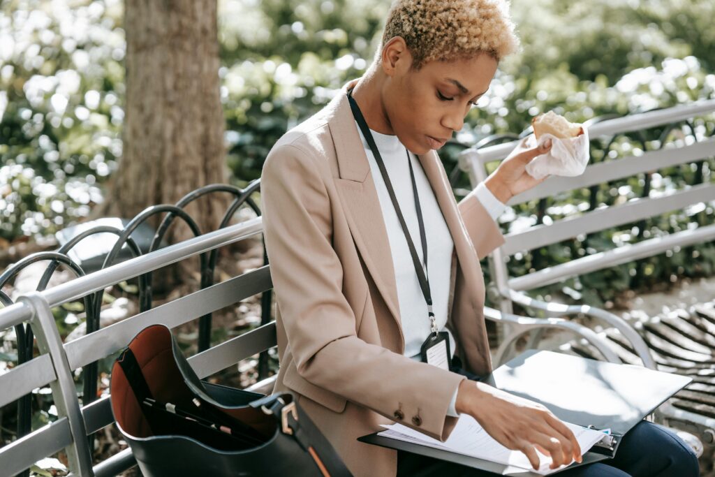 Crop concentrated African American female in classy jacket with badge eating sandwich while reading documents in folder sitting on bench in park